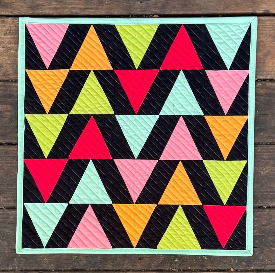 Colorful triangles quilt on black background with light teal binding, photographed on a wooden deck.